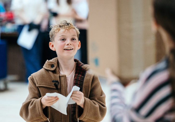 A young boy in a brown trench coat smiles gently. He is holding a folded piece of paper in his hands.