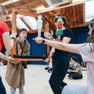 A group of young performers are in a rehearsal studio. Two of them are holding a large wooden frame, which another boy in a beige trench coat is looking through in wonderment. Another person is holding up two nutcrackers and a small wooden reindeer.