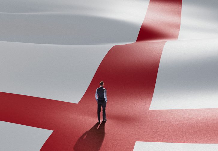 A small figure of a man stands with his back to the camera in the centre of a giant Cross of St George - the England football team's flag. The figure is dressed in Gareth Southgate's signature waistcoat, shirt sleeves and suit trousers.