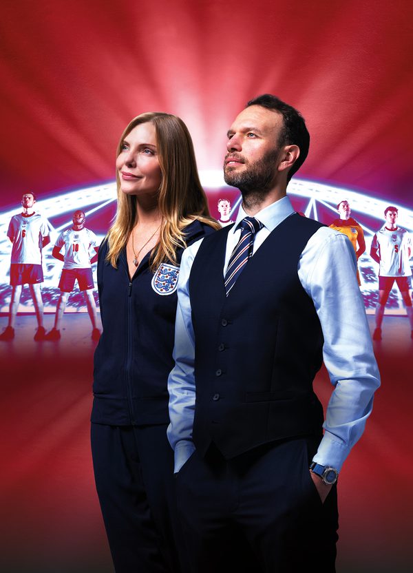 A man and a woman stand beside one another, proudly looking up into the distance. The woman (labelled Samantha Womack as Pippa Grange) wears an England football team tracksuit with the three lions badge. The man (labelled David Sturzaker as Gareth Southgate) wears Southgate's iconic waistcoat over a shirt and tie. The red background features an illuminated stadium with England football players lined up proudly in front of it. Text over the image reads: 'Five stars (The i), 'The decade's biggest theatrical triumph' (Telegraph). The acclaimed National Theatre production: Dear England. A new play by James Graham, directed by Rupert Goold, set design by Es Devlin.'