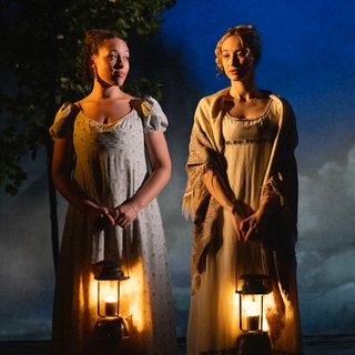 Two young women in Regency dress are standing next to each other, looking pensive. It's an evening scene and both are carrying lighted lanterns.