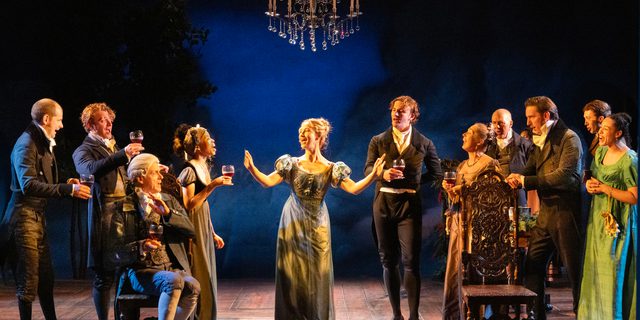 A group of men and women in elegant Regency evening dress are raising a toast to the young blonde woman in the centre, who extends her arms in acknowledgement. A chandelier and ceiling cornice is above them.