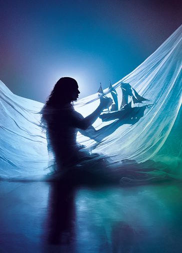 A silhouette of a woman holding a model ship, with a white sheet draped in front of them. They are surrounded by moody green and blue lighting with a haze effect.