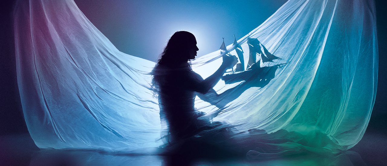 A silhouette of a woman holding a model ship, with a white sheet draped in front of them. They are surrounded by moody green and blue lighting with a haze effect.