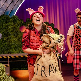 An actor stands at the front of the stage singing. She is wearing all pink and pig ears and a snout. In her arms she is holding a burlap sack with straw written on it. There is straw spilling out of the bag. Behind her, another of the Little Pigs, wearing a pink leopard print vest and pink tartan shorts, is looking at her in amazement.