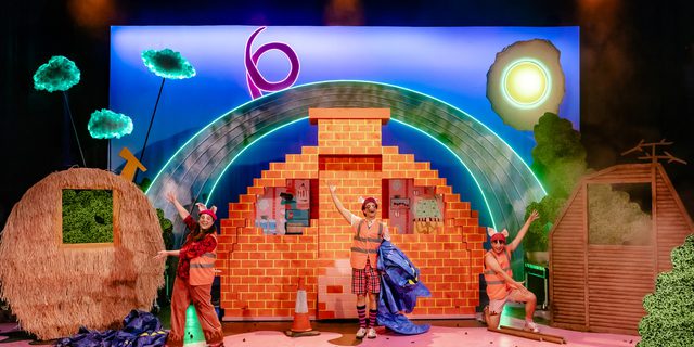 Three actors perform on a brightly lit stage. They wear playful pink and patterned costumes with pig ears and snouts. On the left, there is a straw house, in the middle, one made of bricks and on the right, a house made of sticks. They are smiling and singing in front of a large brick house set piece. Behind them, there is a colourful neon rainbow and clouds.