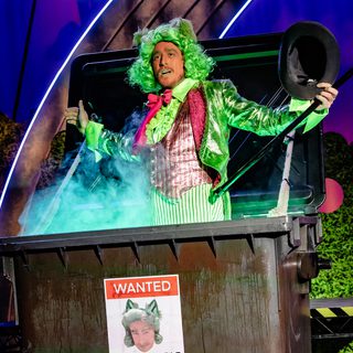 A man in pinstripe trousers and a matching waistcoat, with a metallic jacket and a green curly wig with pointy ears is stood in an industrial bin which has a wanted poster with his face on plastered across the front. He is singing and holding a top hat and cane.