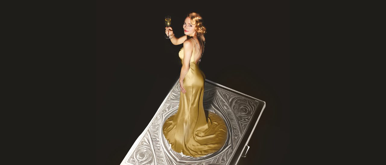 A giant, decorative, silver cigarette case floats against a black background. A young woman stands on top wearing a glamorous golden silk gown, holding a glass of champagne and looking to the camera with a smile. Her golden hair is curled and she wears red lipstick.