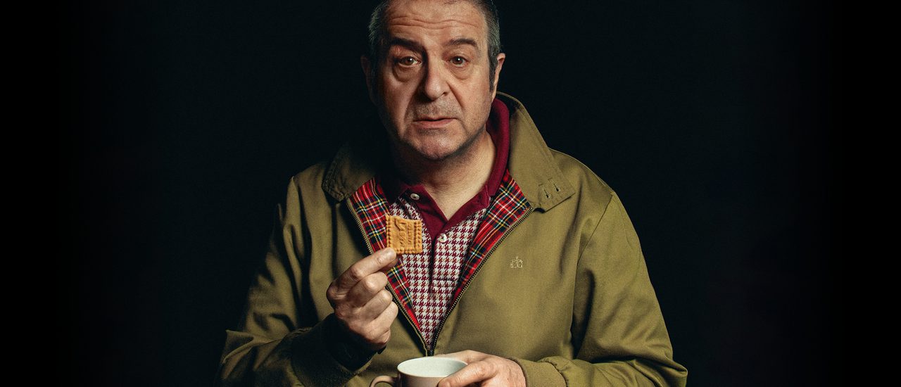 A man with short grey hair holds a biscuit and a cup of tea, looking out with a tired, vacant expression. He wears a khaki jacket and checked burgundy and white polo shirt.