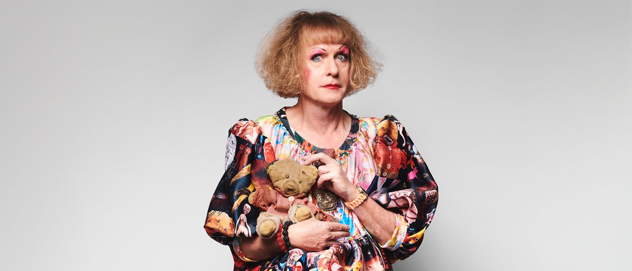 Grayson Perry, a man with long dark blonde curly hair and a fringe, wears a brightly coloured, patterned dress with puffy sleeves and exaggerated make-up, holds a small teddy bear. He is wide-eyed.