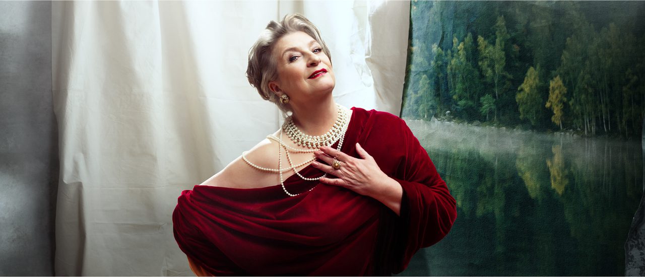 An older woman leans to one side, endearingly looking at the camera with a slight smile. She is heavily made up with dark red lipstick and has her grey hair tied back. She wears a luxurious dark red velvet one-shoulder dress, layers of pearls, a large ring and big round earrings.