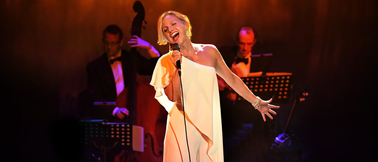 A blonde woman wearing a glamorous, white, one-shoulder dress sings passionately into a microphone with the other arm extended out to the side. Two musicians can be seen behind her, lit in orange and pink.