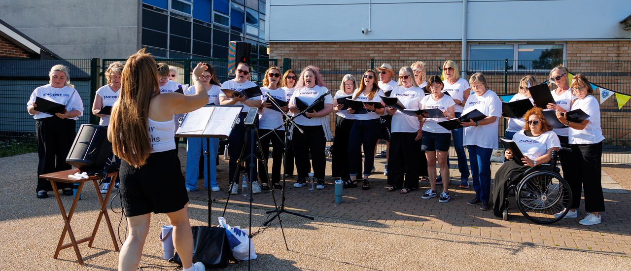 A choir sing together wearing white t-shirts and black bottoms. Their conductor stands in front of them with a music stand, arms outstretched. They are outside with buildings behind them. The sun is shining.