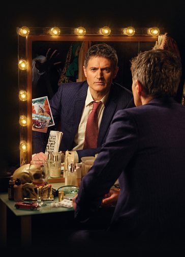 A middle-aged man with short, grey hair sits in front of a dressing table, looking into a mirror surrounded by lights. He is photographed from behind, revealing his reflection. He has an intense stare and a frown. He wears a blue pin -stripe suit jacket, white shirt and red patterned tie and holds a folded newspaper. The dressing table is cluttered with an array of objects, including a nearly-empty wine glass, skull, make-up, pills and toiletries.