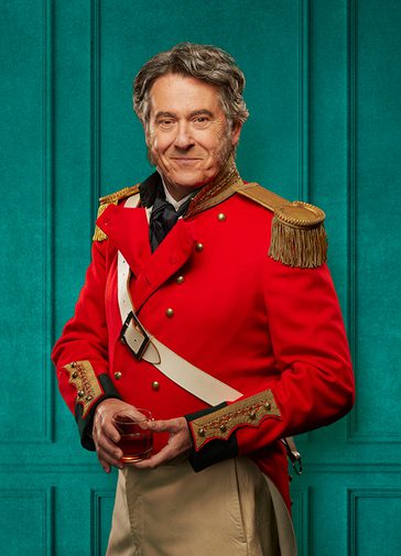 An older man with grey hair and exaggerated side burns stands proudly wearing ceremonial military full dress, including beige trousers and a red jacket with golden buttons, cuffs and epaulettes. He holds a glass of whiskey and looks straight down the camera with a knowing smile.