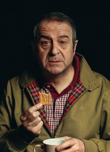A man with short grey hair holds a biscuit and a cup of tea, looking out with a tired, vacant expression. He wears a khaki jacket and checked burgundy and white polo shirt.
