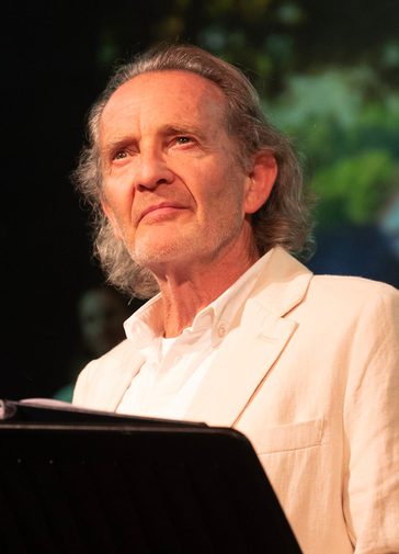 Anton Lesser, an older man with long grey hair, stands behind a lectern, wistfully looking out.