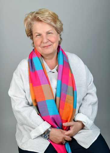 Sandi Toksvig, a woman with short blonde hair, smiles warmly, wearing a white shirt and colourful pink, purple and blue checked scarf.