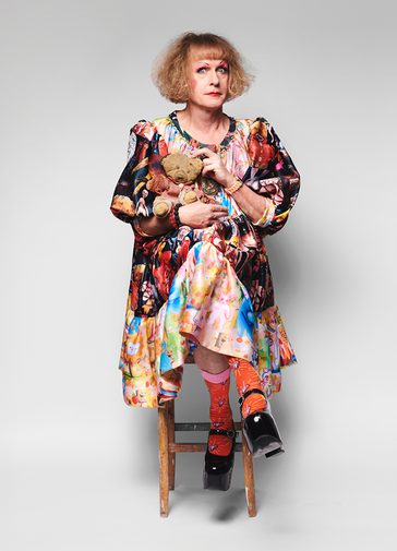 Grayson Perry, a man with long dark blonde curly hair and a fringe, wears a brightly coloured, patterned dress with puffy sleeves and exaggerated make-up, holds a small teddy bear. He is wide-eyed.