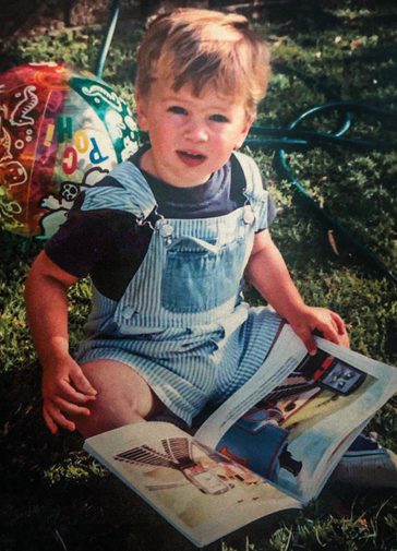 A vintage photograph of a young boy sitting in the grass, holding a magazine with a colourful beach ball behind him. He wears a dark blue t-shirt and blue and white stripey dungarees.