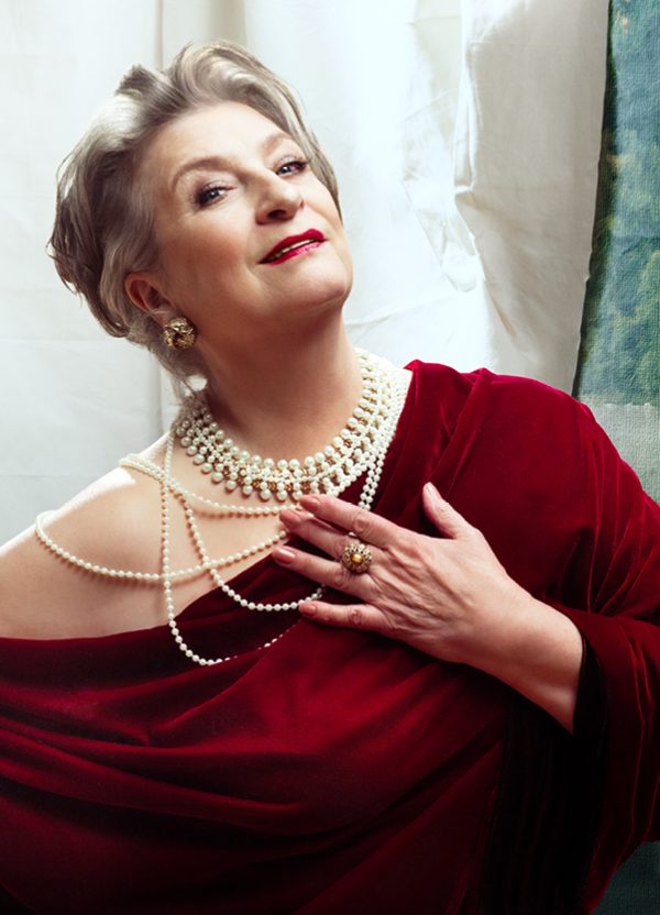 An older woman leans to one side, endearingly looking at the camera with a slight smile. She is heavily made up with dark red lipstick and has her grey hair tied back. She wears a luxurious dark red velvet one-shoulder dress, layers of pearls, a large ring and big round earrings.
