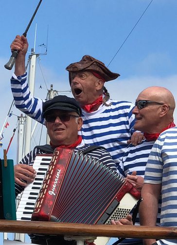 Three men wearing white and blue stripey tops with red neckerchiefs. One plays an accordion whilst another looks out to the side, open mouthed, exclaiming happily.