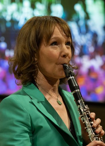 Emma Johnson MBE, a woman with mid length brown hair wearing a green blazer, plays a clarinet. In the background is a colourful graphic projection with shades of blue, purple and green.