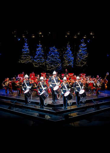 The Band of HM Royal Marines stand on the Festival Theatre stage dressed in military full dress, facing out front. Five drummers stand at the front, with the rest of the band seated behind. Behind them are five blue-lit Christmas Trees.
