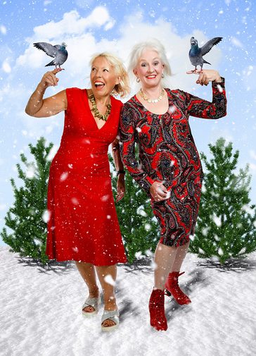 Two older ladies stand shoulder to shoulder, holding birds on their fingers. The one on the left wears a bright red dress and the one on the left wears a red paisley printed dress. There are green Christmas trees and a cloudy sky behind them. They look glamorous, very happy and jolly.