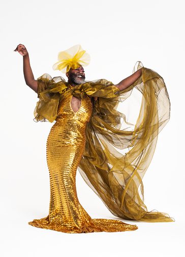 A Black drag performer with a big grey beard wears a dazzling, gold, sequined ball gown with puffy tulle sleeves, an exaggerated train and a yellow flower headpiece. They smile and hold both arms out in a sassy flourish.