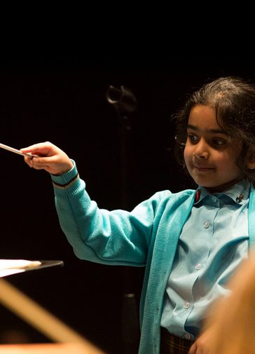 A young girl is smiling as she waves a conductor's baton at an orchestra. In front of her is a music stand. She wears a blue school uniform.