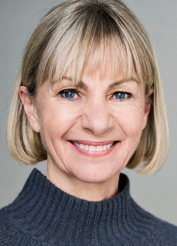 Kate Mosse, a woman with a neat blonde bob, smiles warmly. She wears a dark grey, turtle-neck jumper.