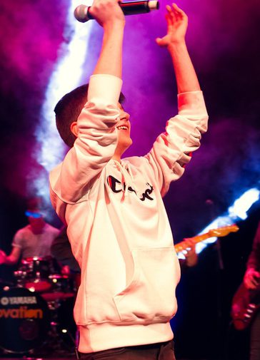 A boy in a white sweatshirt holds his arms above his head, holding a microphone. Several spotlit musicians are behind him.