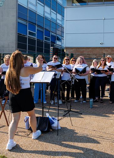 A choir sing together wearing white t-shirts and black bottoms. Their conductor stands in front of them with a music stand, arms outstretched. They are outside with buildings behind them. The sun is shining.