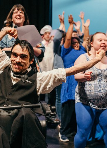 A group of learning-disabled and neuro-divergent people performing, dancing and laughing. In the forefront, a male wheelchair user, wearing a white shirt and black waistcoat, enthusiastically waves a piece of paper above his head, his other arm spread out wide.