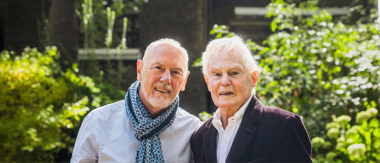 Two men (Derek Jacobi, right, and Richard Clifford, left) are stood side by side, an arm around each other. The man on the right (Derek Jacobi) is wearing a purple courdroy blazer and a white shirt, the man on the left (Richard Clifford) is wearing a blue striped shirt and a blue patterned scarf. They are both smiling at the camera.