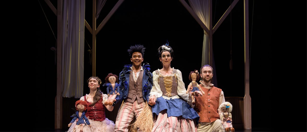 Four members of the company stand holding hands and smiling. They are holding puppet versions of themselves. They are all dressed eccentrically, with mismatched outfits. The man and woman in the middle are taller than the others. They both have their eyebrows raised.