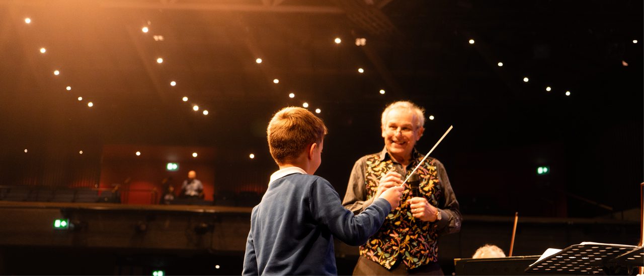 A small boy with his back to the camera stands holding a conductor's baton. An older man, wearing an embroidered waistcoat, stands smiling down at him. They are on stage with music stands beside them.