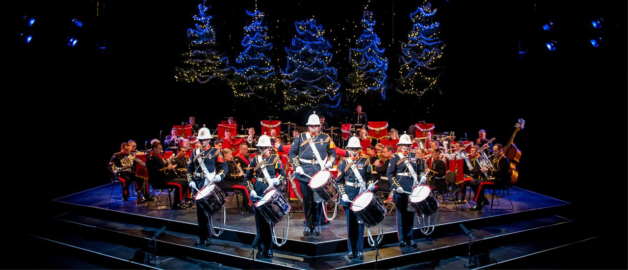 The Band of HM Royal Marines stand on the Festival Theatre stage dressed in military full dress, facing out front. Five drummers stand at the front, with the rest of the band seated behind. Behind them are five blue-lit