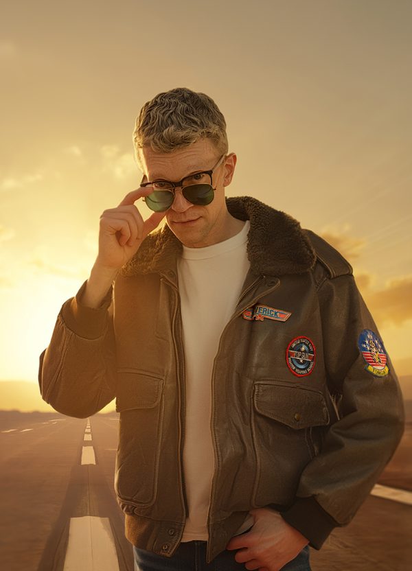 A spoof of an image from the iconic film Top Gun: a man stands wearing a leather aviator jacket, white t-shirt, blue jeans, glasses with thick, black frames, and aviator sunglasses. He holds the sunglasses lower down on the bridge of his nose and looks over them at the camera with an inquisitive facial expression. Behind him is a road and a dusty, golden sky.