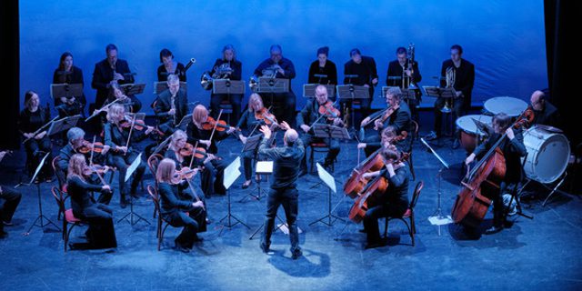 A full orchestra play their instruments. A conductors stands in the middle, his hands raised up with his conducting stick. They are all dressed in concert blacks and a blue wash covers the stage.