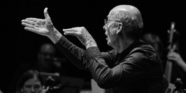 A black and white image of a conductor in a shirt and smart trousers. His hands are raised, conducting. A few of the orchestra are playing their instruments in the background.
