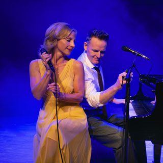A man and woman sit on a piano stool. The man is playing the piano in a white shirt with the sleeves rolled up and a black tie, a microphone stand next to him. He is looking at the side to the woman who is sitting on the edge of the seat. She is wearing a long yellow sleeveless dress and is holding a wired microphone up. She is also looking to the side. A blue wash of lights behind them.