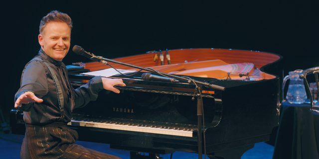 A man sits at a grand piano. He is wearing a black shirt and braces and black trousers. He is turned away from the piano, a wide smile on his face and an arm outstretched in front of him, gesturing.
