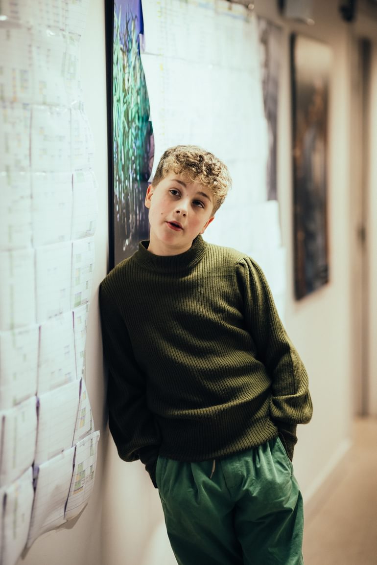 A young performer wearing a green jumper and green trousers leans against a white wall, mid conversation. Their hands are in their pockets and they look relaxed and casual.