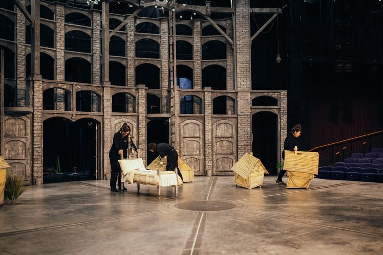 Three members of stage crew, wearing all black work on stage, prepare the set on stage. The backdrop is a greyish brown brick wall with arched windows. The floor is grey stone cobbles. One wheels on a grey bed dressed with cream bedding. The other two wheel on houses, in the style of paper lanterns. The tiered theatre seating and stage lights can be seen in the background on the right hand side of the image.