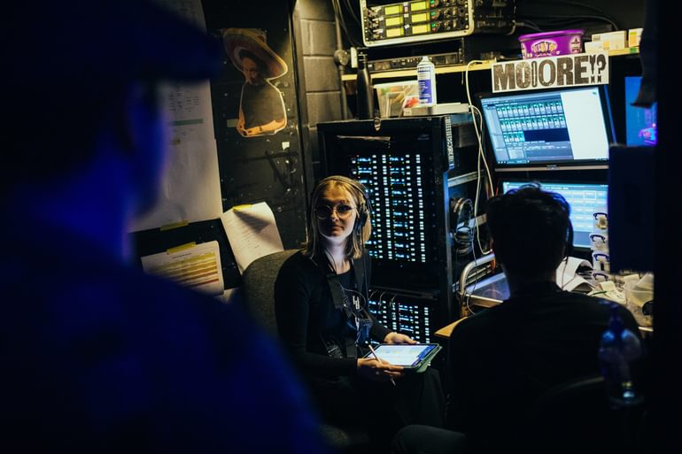 A young person wearing all black holds a tablet and sits backstage in front of two screens and a radio mic rac. They wear a headset and look at a performer, out of focus in the foreground, focused and friendly.