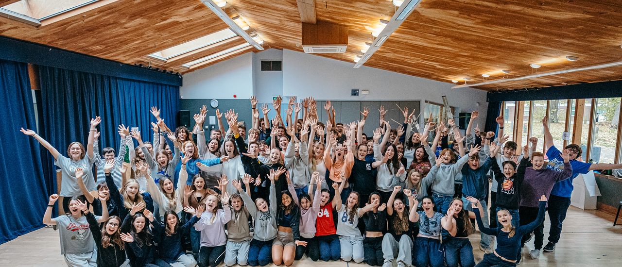 A huge group of young performers fill a rehearsal room. They stand and sit close together, posing with their arms spread wide in the air and big cheesy smiles. They are all very happy.