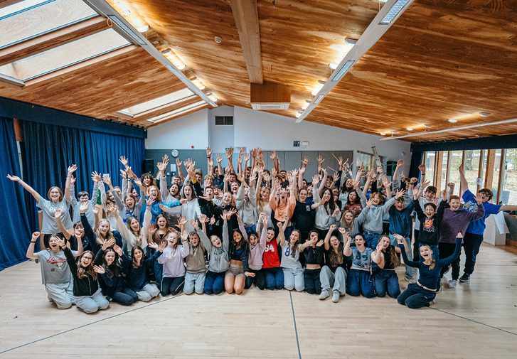 A huge group of young performers fill a rehearsal room. They stand and sit close together, posing with their arms spread wide in the air and big cheesy smiles. They are all very happy.
