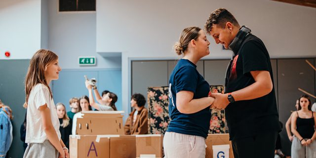 Two young performers clutch hands and stand close together, their foreheads very nearly touching. They look in love. A smaller girl watches on behind them and smiles. In the background other young performers watch, out of focus.